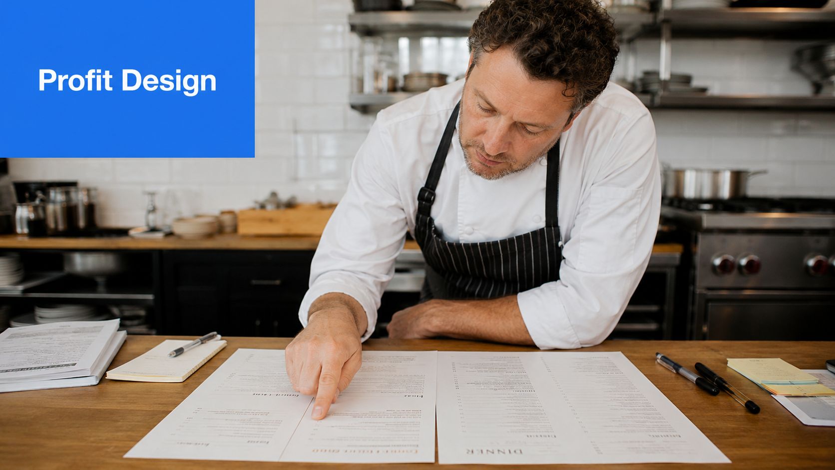 A professional chef pointing at a paper menu while standing in a busy commercial restaurant kitchen.